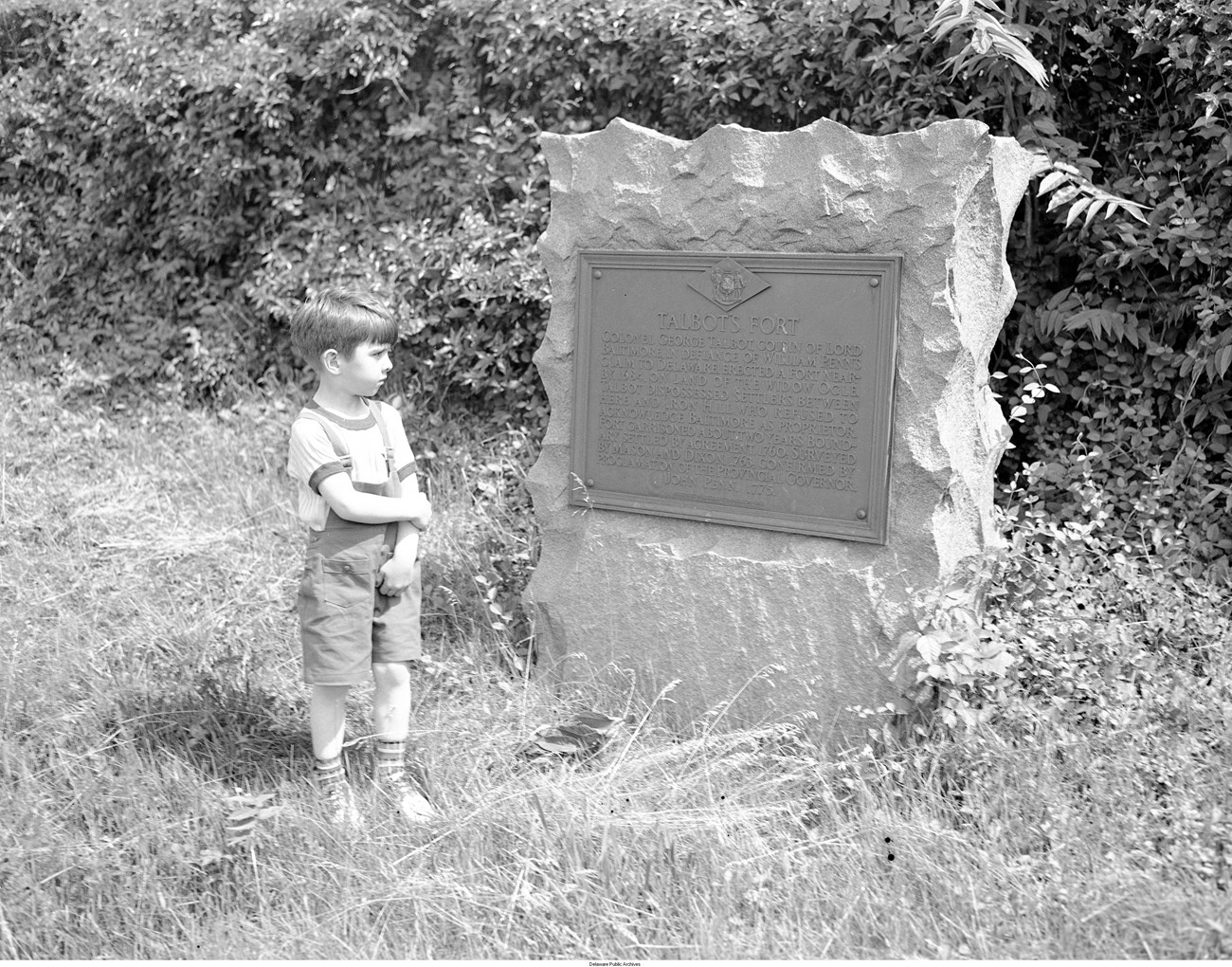 Fort Talbot Marker Young boy wearing overall shorts stands next to a large stone with historical plaque about Talbot’s Fort.
