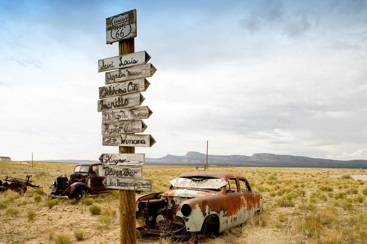 Two rusted out cars in a open field next to a post with arrows pointed to different city on route 66.
