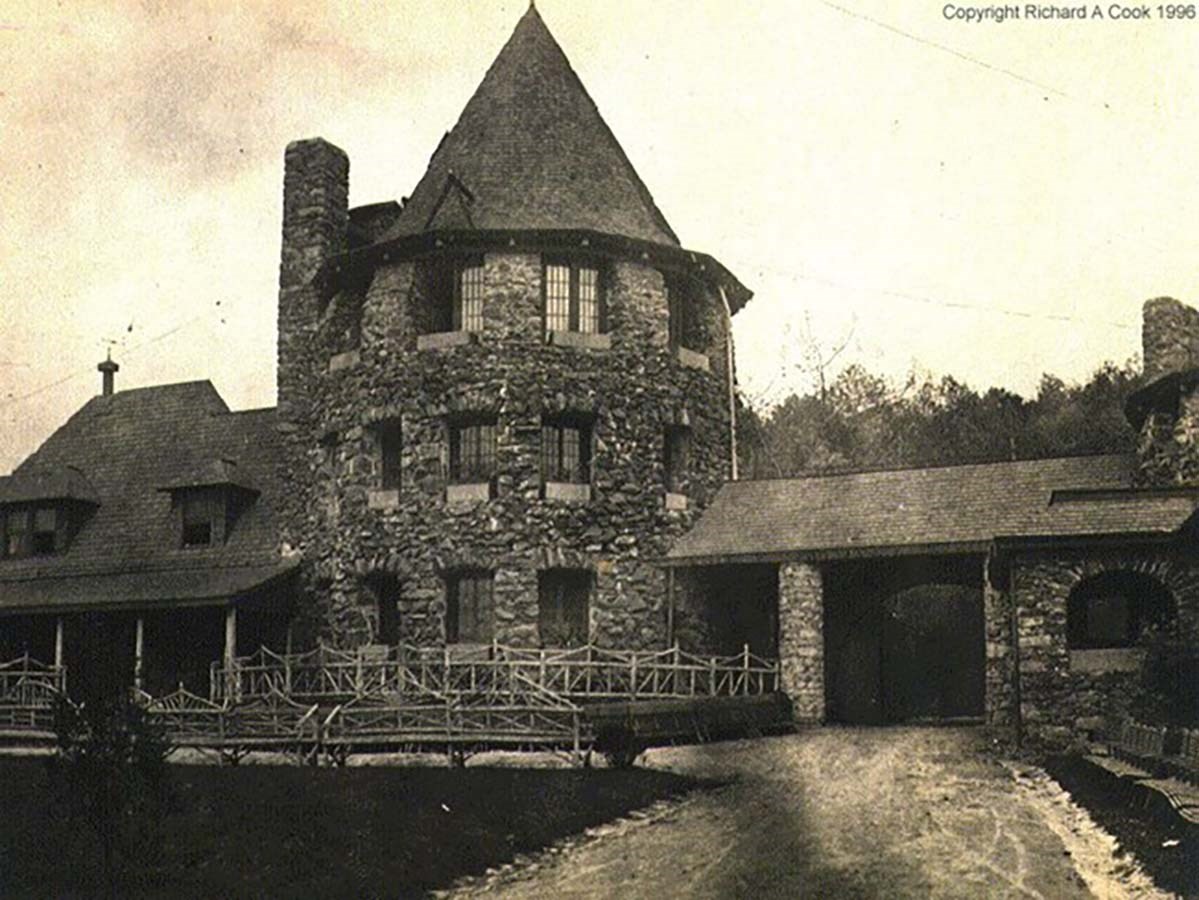 Historic photo of three-story stone turret tower attached to a house on the left, and a driveway and garage on right.