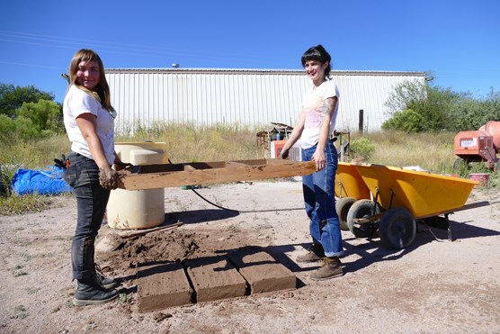 lt= two people hold a wooden form above newly made adobe bricks