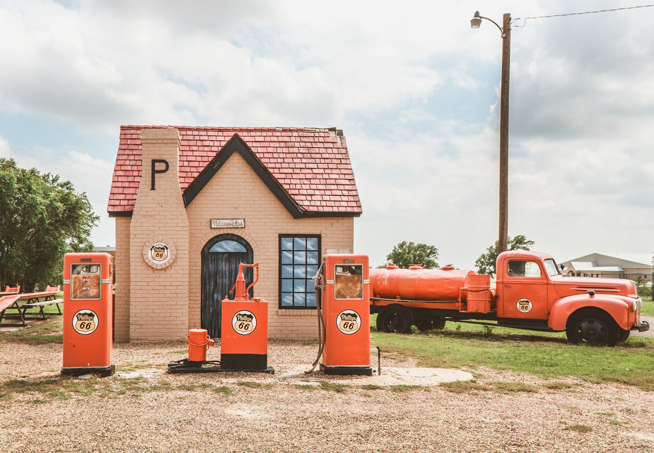 3 refurbished orange gas pumps stand in front of a peach brick building and an orange gas truck park next to it.
