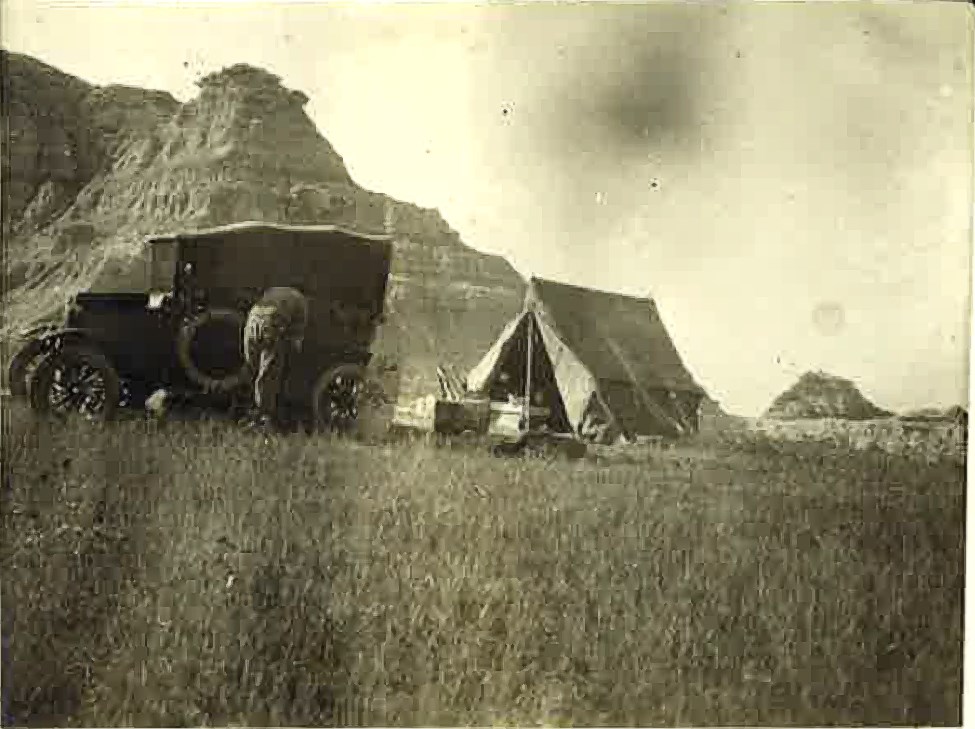 historic photo showing a person, a car and a tent in a field with badlands formations in the distance