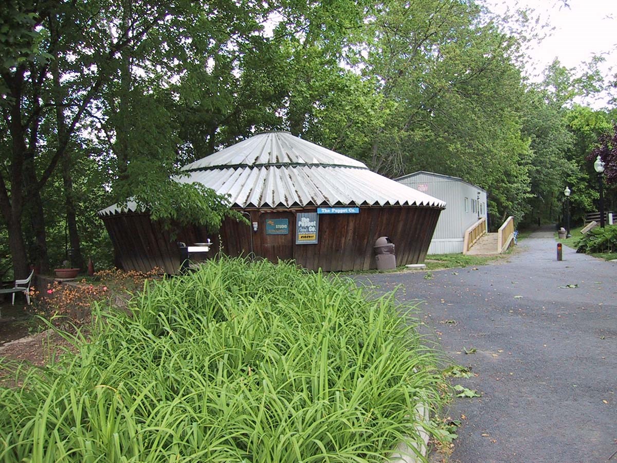 A wide black walkway leads to a brown wooden circular yurt with silver metal roof surrounded, trees in background.