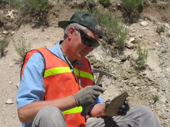 a person sitting on the ground tapping a rock with a rock hammer