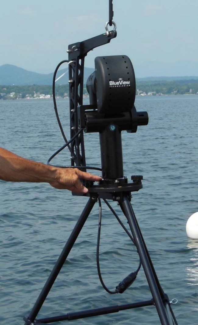 A tripod mounted device labeled BlueView on a boat, water in the background, a hand with finger pointing to the tripod.
