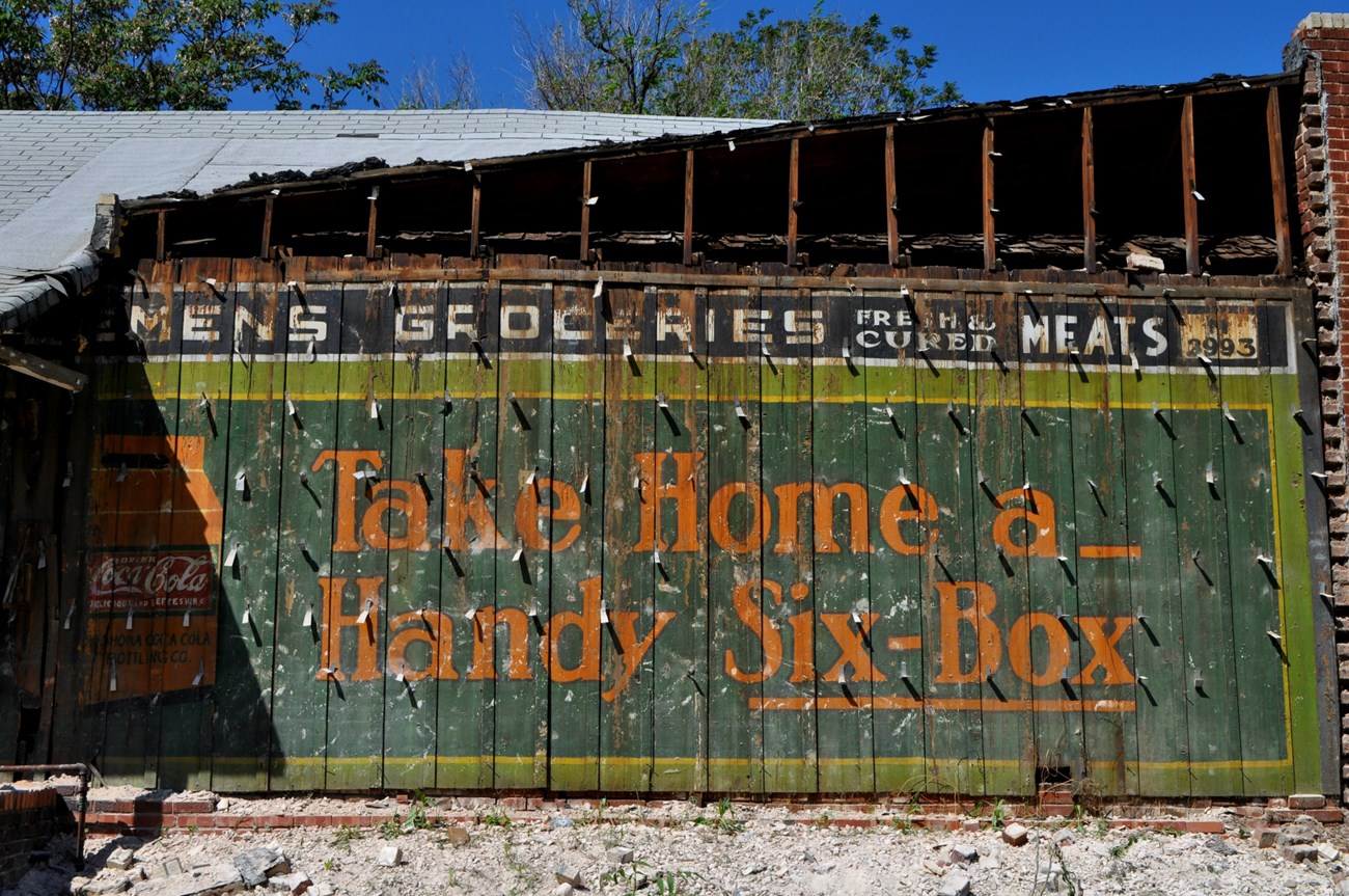 Coca-Cola Advertisement Weathered sign on a greenwood plank wall with Coca-Cola logo and orange text reading Take Home a handy six-box.