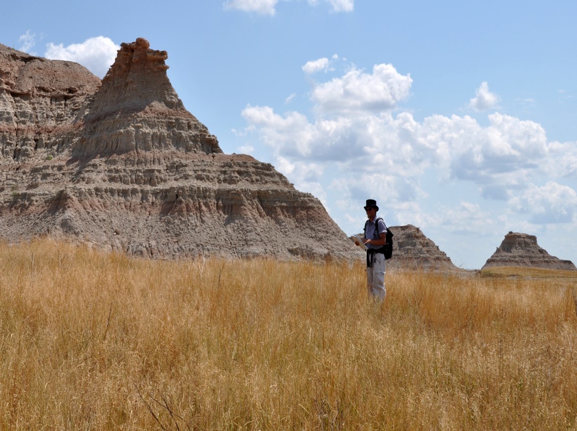 photo of a person standing in field of dry grass with badlands formations in the distance