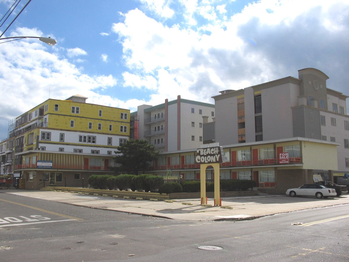 For Sale Two story weathered L shaped motel building in front of modern buildings with sign reading Beach Colony.