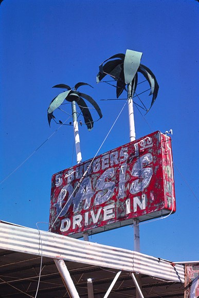 Oasis Drive In Weathered neon sign two fake palm trees and red rectangle with white text reading Oasis Drive-in.