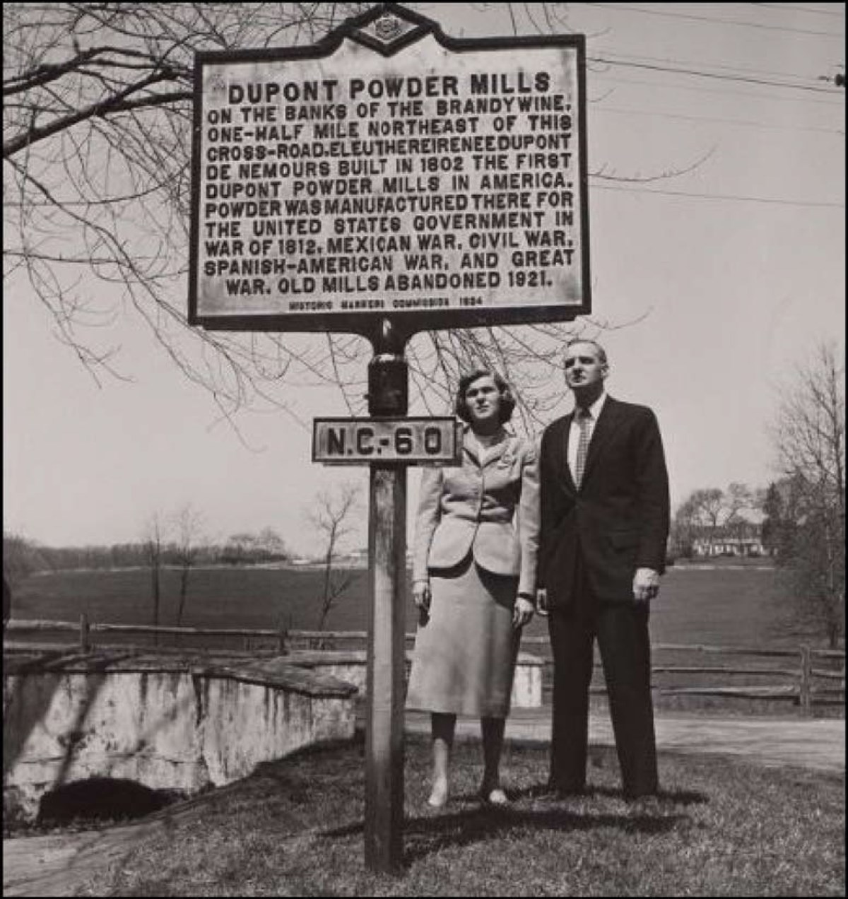Man and woman reading Dupont Powder Mills historic roadside marker N.C. – 60. Countryside in background.