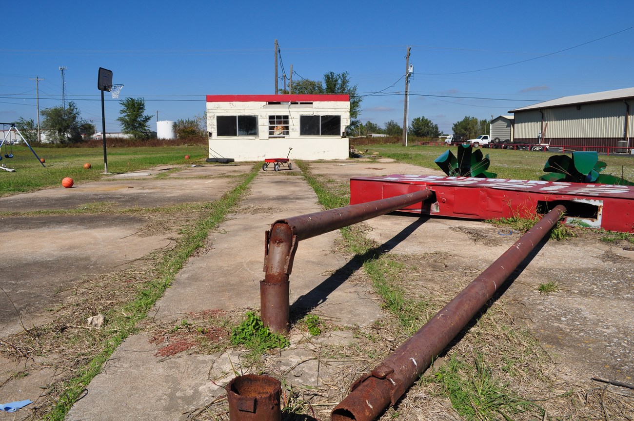 Collapsed sign. Grass growing in cement lot with small whit building in background. Foreground rusted poles of a collapsed sign.