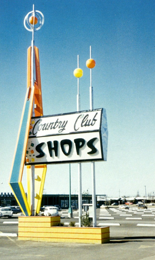Country Club Shops sign. Yellow base left polygon rises to orb encased in circles right two poles with yellow and orange orbs.