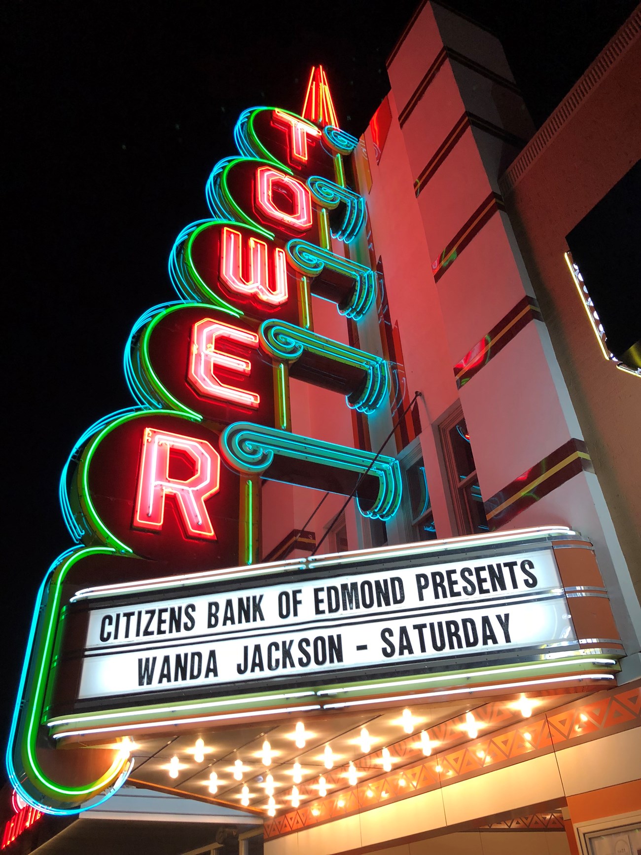 Brightly lit in pink neon vertical text Tower with green and turquoise trim and white marquee.