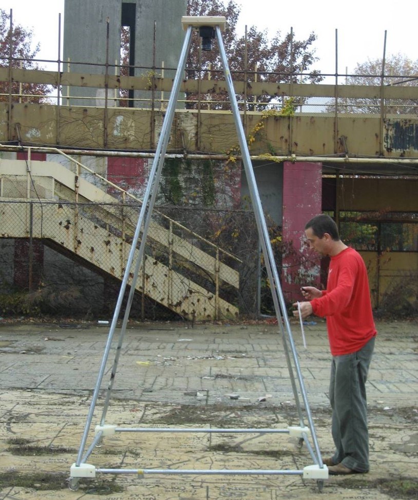 Man in red shirt standing next to tall rolling pyramid with camera at top pointing toward the ground.