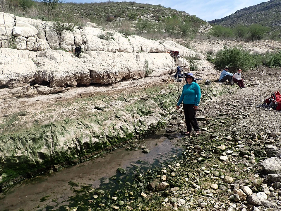 Indian Springs Canyon Spring Two people sitting and one person standing on the edge of a stream pointing at the orifice of the spring that emerges as moderate flow in a stream channel lined with cobbles. Two limestone ledges are seen along one side of the stream.
