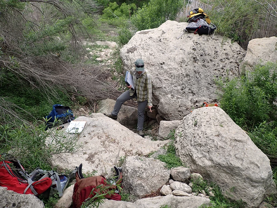 Mouth of the Pecos Spring A person points to a dry area on the ground surrounded by large gray boulders and bright green vegetation.
