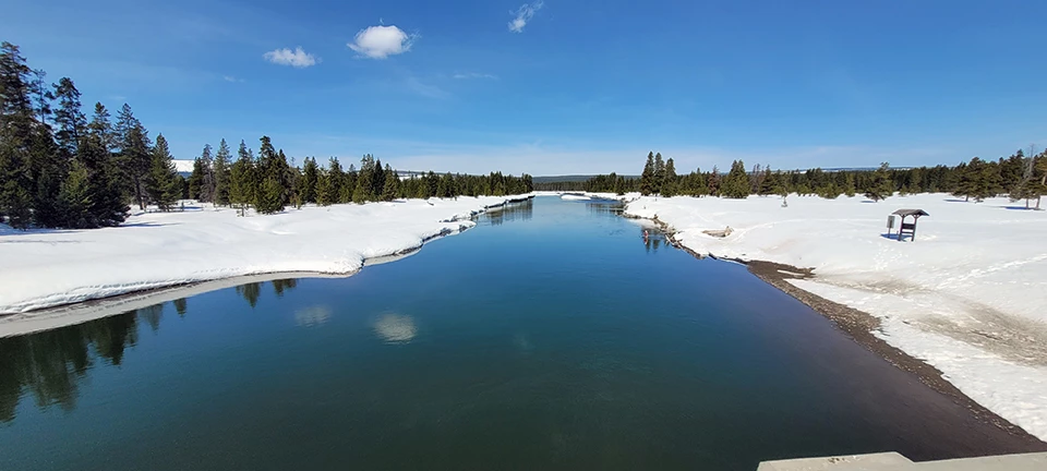 Snake River at Flagg Ranch, Wyoming A calm, blue river with mostly flat, snowy banks in a snowy landscape of scattered conifers under a deep blue sky.