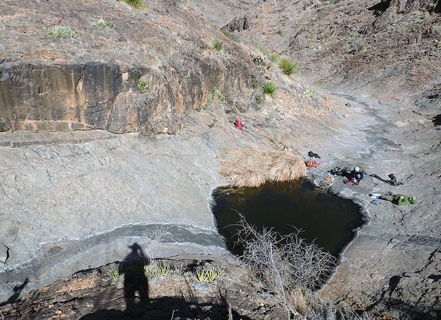 Scientists near a giant pool of water in a bedrock basin in a steep, rock-lined drainage and evidence (water marks) that water flows from above into the pool and out from the pool down the drainage.