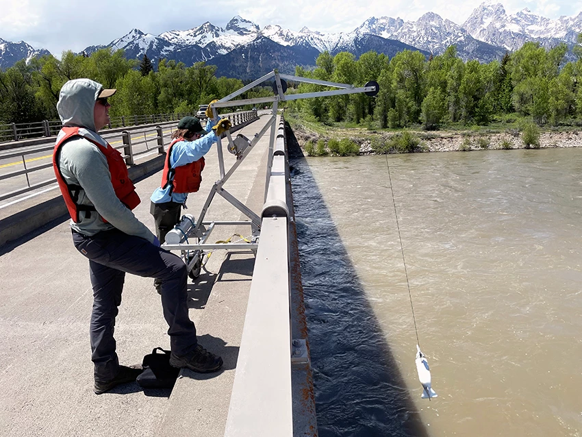 Water sampling on the Snake River Two scientists standing on a bridge over a river turning a crank on a crane to lower a reel with equipment for collecting water samples into the river.