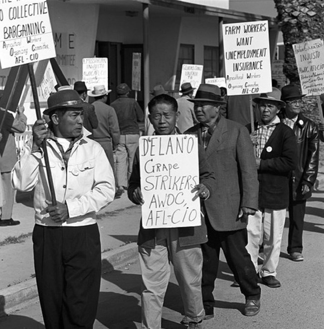 Several men walk in a line carrying AWOC picket signs in front of a building Several men walk in a line carrying AWOC picket signs in front of a building