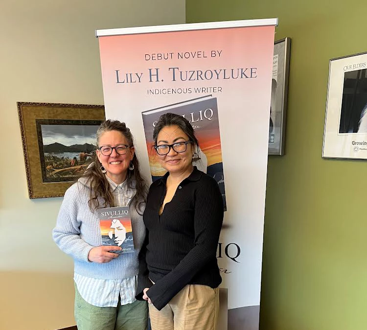 Inupiaq writer Lily H. Tuzroyluke and Lisa Fink Two women standing in front of a sign that says, "debut novel by Lily H Tuzroyluke"