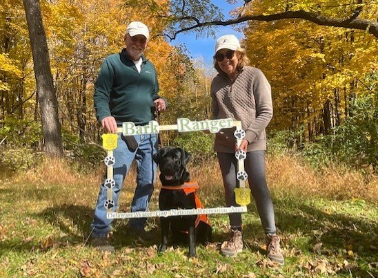 A black dog in an orange vest sits in between his owners framed by a photo op picture frame.