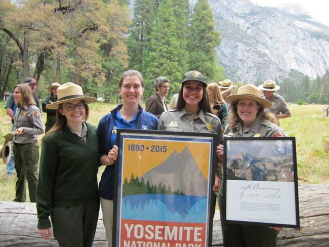 Four people in natural landscape holding posters related to the National Park Service Centennial in Yosemite National Park.