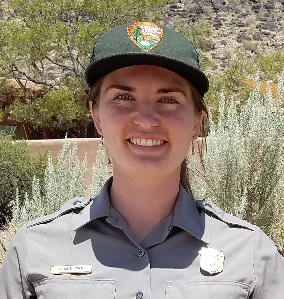 Young female wearing the NPS uniform with a ball cap and NPS logo in the center. There are green trees, rocks, and a blue-white sky behind her.