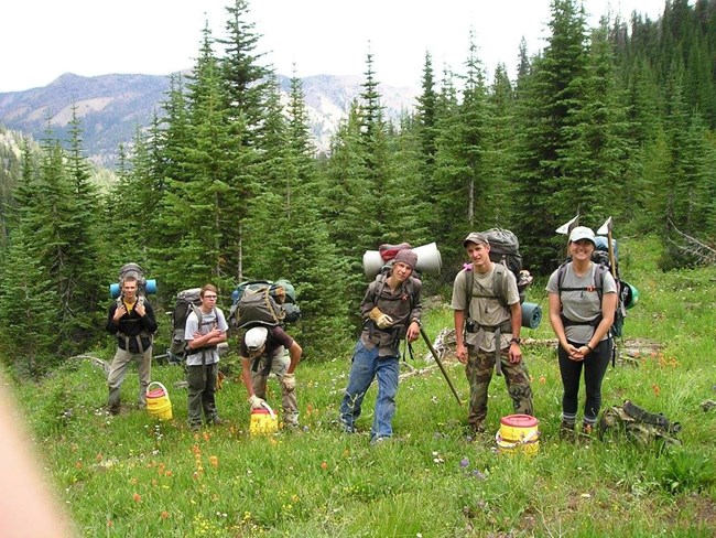 Six youth corps members wearing backpacks and carrying tools on their way to a backcountry trip. There are green trees, grass, and greyish sky in the background.