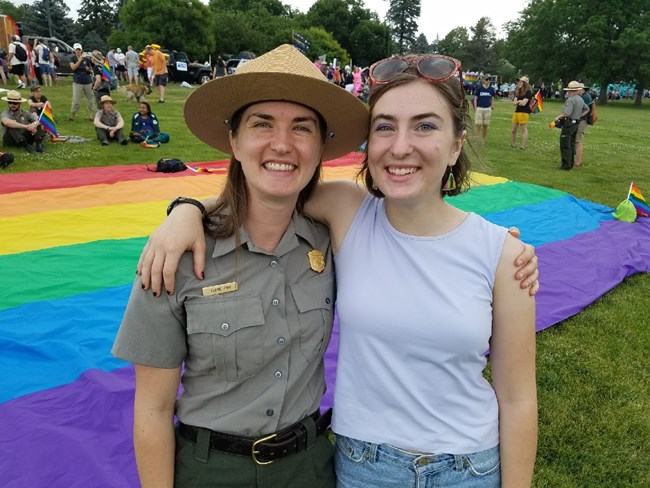 Claire and her sister at the Denver PrideFest Parade Two young females stand in front of a pride flag with other people in the background. The one on the left is in the NPS uniform; the other is wearing a tank top and jeans.