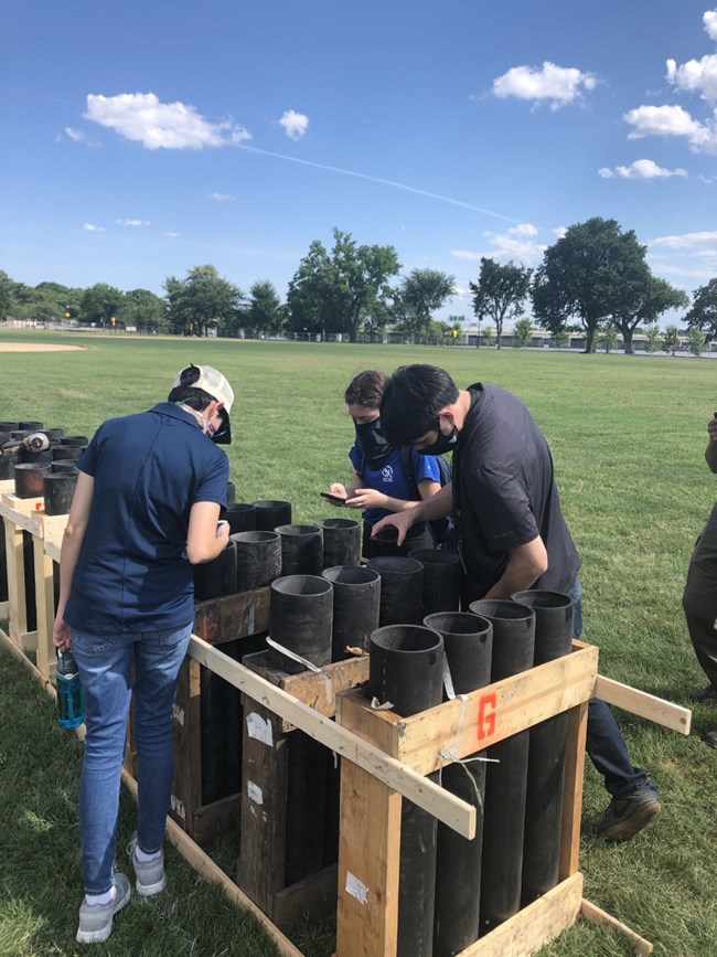 National Mall Fireworks Inspection People lean over to look inside large plastic tubes placed in wooden racks.