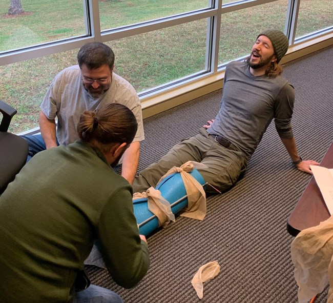 Man laying on the floor with his leg wrapped two other people kneeling beside him