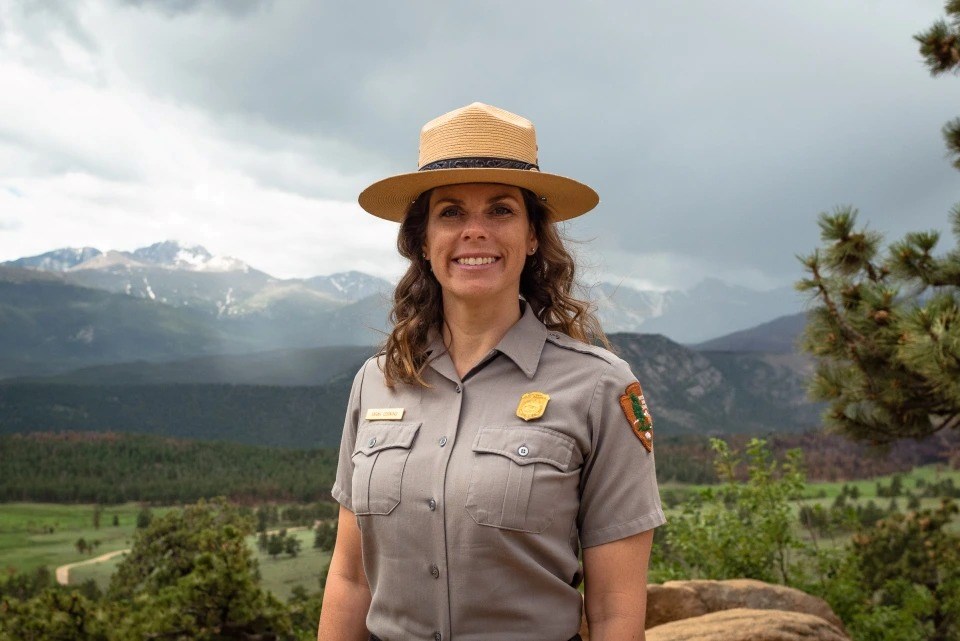 A female in a National Park Service uniform smiles against the background of Rocky Mountain National Park.