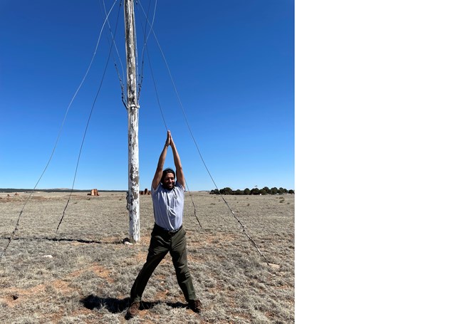 Park Ranger standing erect with arms raised above their head and hands meeting together, with flagpole in background