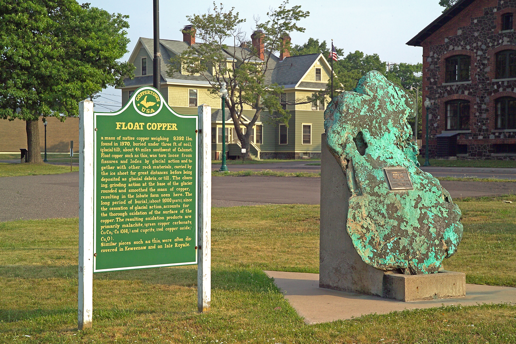 A large, piece of mass copper with a blue-green patina next to a green sign with the title: "Float Copper" make up the foreground. A stone building and wooden structure are in the background with green leaved trees completing the landscape.