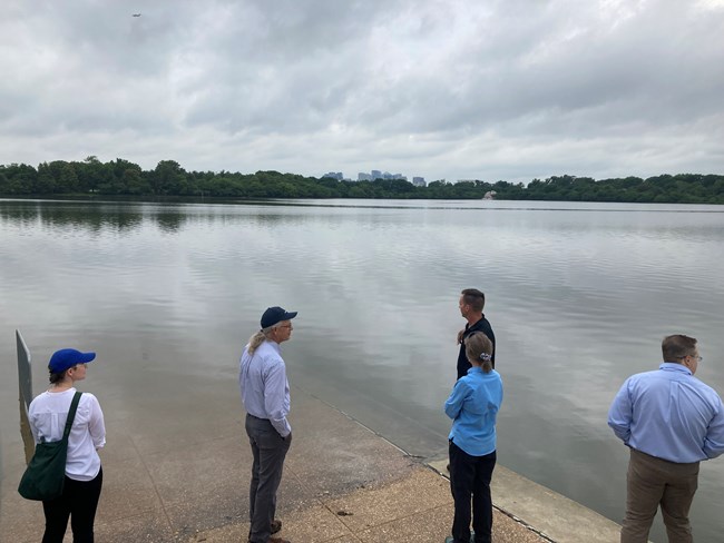 Standing water flooding foreground areas with four people in business attire standing on the water's edge on an overcast day with a city in the distance.