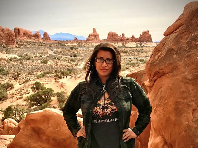 Woman stands in amidst red sandstone rock formations with view of more red rock desert behind her.