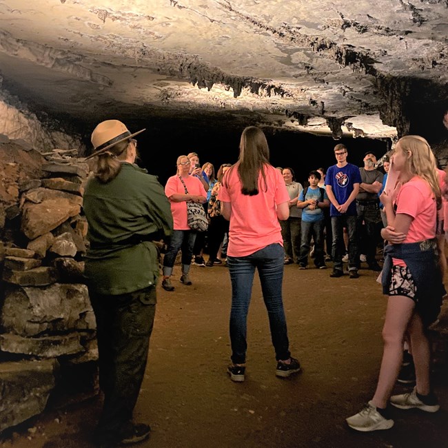Friends and family join the Following Their Footsteps campers on a tour of Gothic Avenue. A group of people stand in a cave with stalagtites and listen to a person speak.
