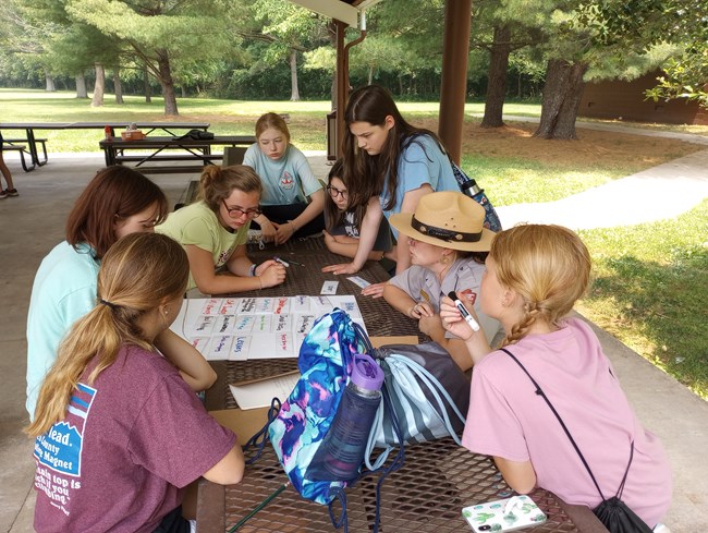 Ranger Lili helps campers record the sights and sounds of Abraham Lincoln Birthplace for an exercise in writing poetry. A group of young women sit around a picnic table and look at a white display board with different color words written on it.
