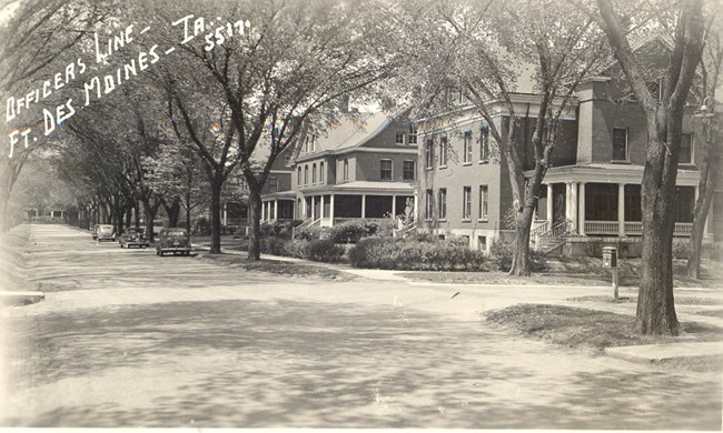 View of stately officers' housing looking down a street. Houses are multi-story with wrap-around porches. Trees line a grassy easement and three cars are along the curb.