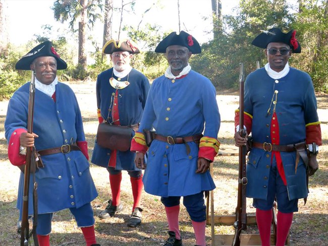 Three African American soldiers in blue uniforms and black tricorn hats smile at camera