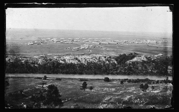 in black and white, across a river, the barracks and command buildings of a military fort can be seen on a prairie