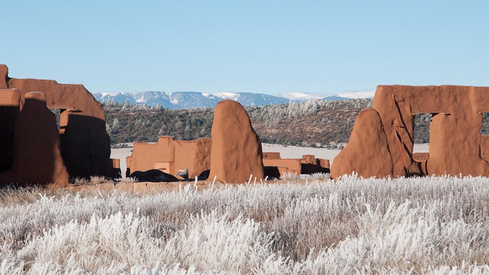 Brown mud ruin walls in a valley of snow covered grasses with snow covered mountains behind them.