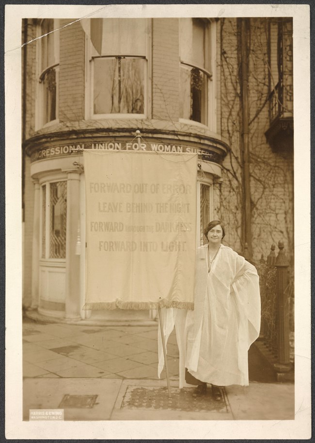 Jessie Hardy MacKaye with banner in front of building. Banner reads: Forward Out of Error Leave Behind the Night Forward through the Darkness Forward Into Light."