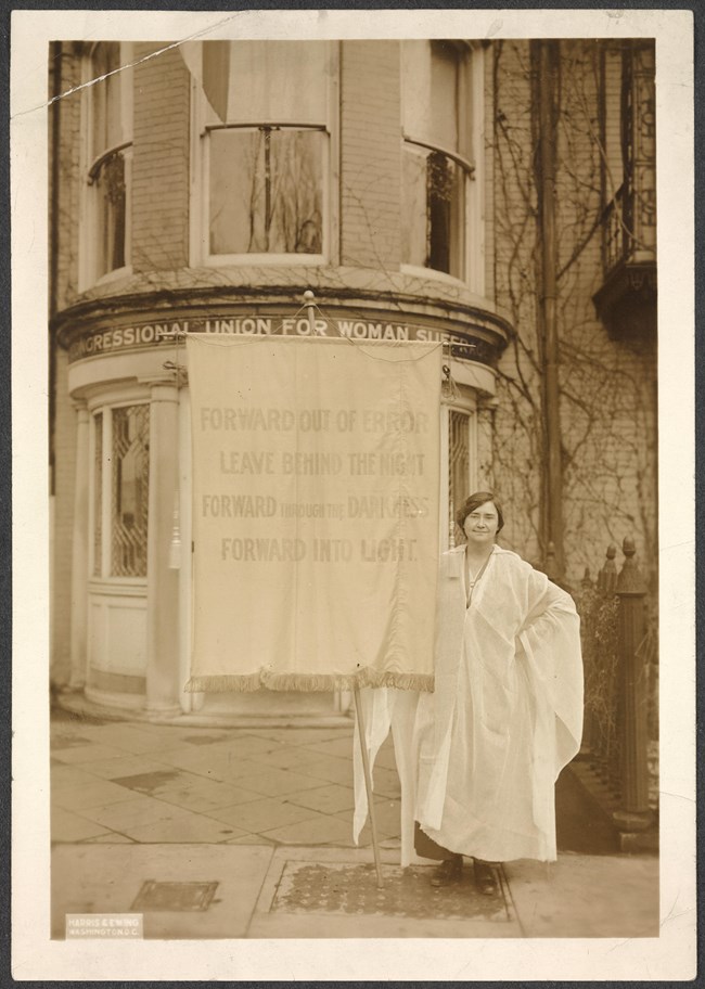 Photograph of Jessie Hardy MacKaye with banner in front of Congressional Union for Woman Suffrage building. Banner reads: Forward Out of Error Leave Behind the Night Forward through the Darkness Forward Into Light." Jessie Hardy MacKaye with banner in front of building. Banner reads: Forward Out of Error Leave Behind the Night Forward through the Darkness Forward Into Light."