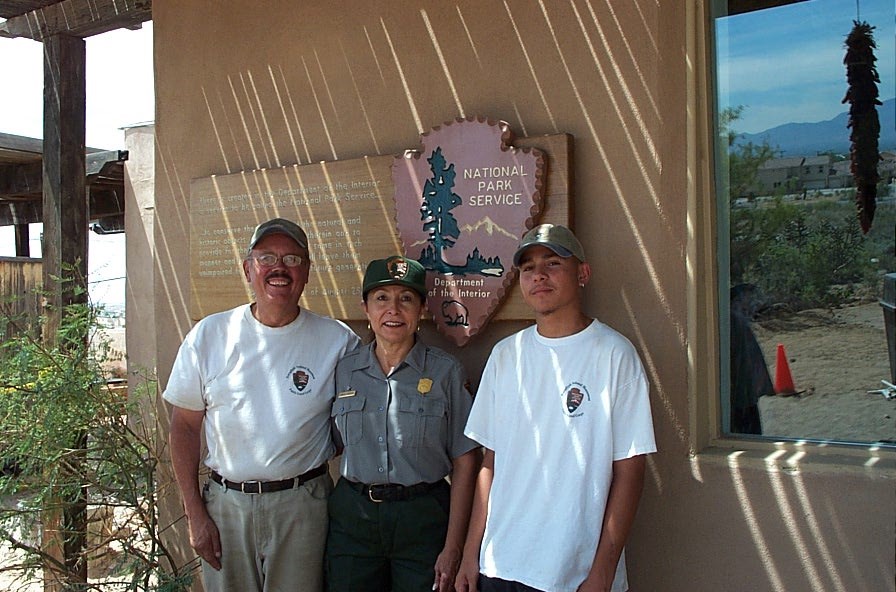 Judith Cordova and members of Youth Development, Inc. Judith Cordova in her NPS uniform poses in between a man and a teenager wearing white t-shirts. They stand in front of an NPS sign hung on an adobe building.