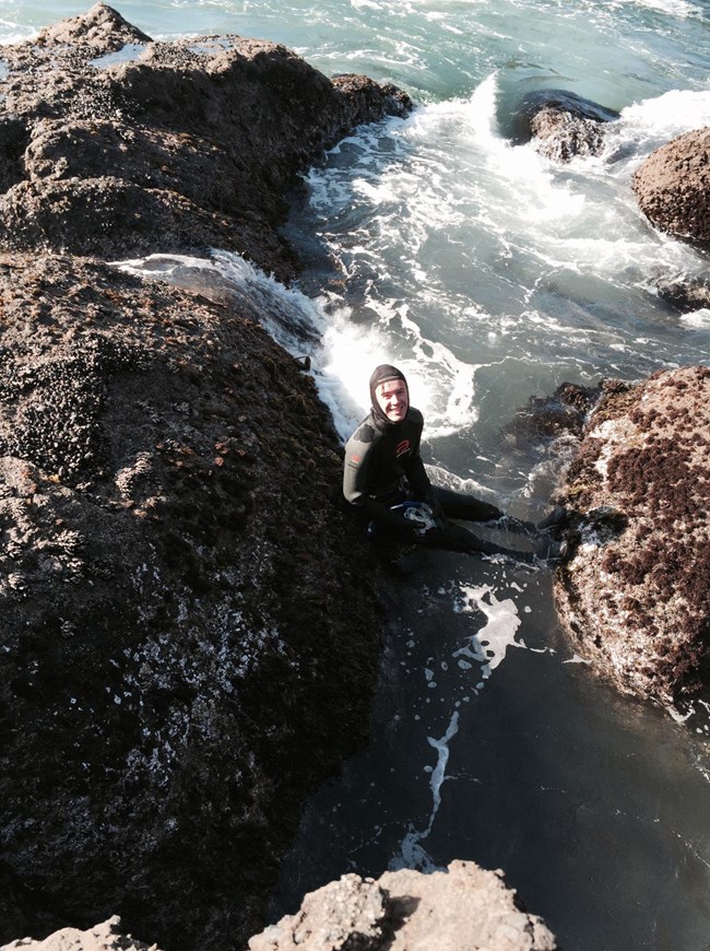 Person in a wetsuit smiles up at the camera while swimming in the ocean between two large rocks.