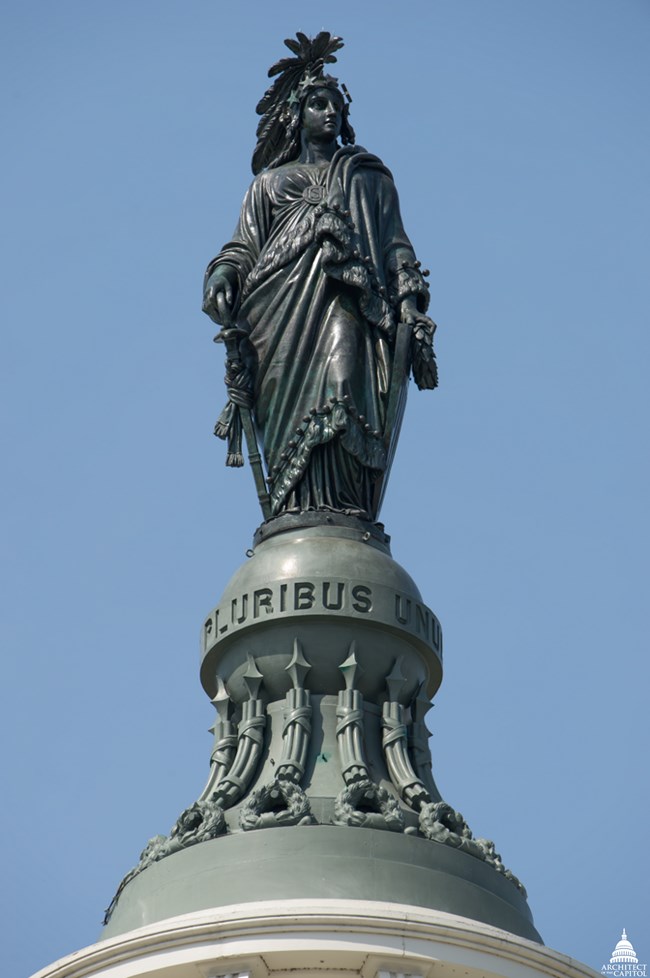 A color photo of the bronze statue of Freedom at the top of the US Capitol dome. Freedom is portrayed as a woman dressed in a Roman toga, holding a sword at her right side and a shield at her left. On her head is a headdress of eagle feathers and stars. T