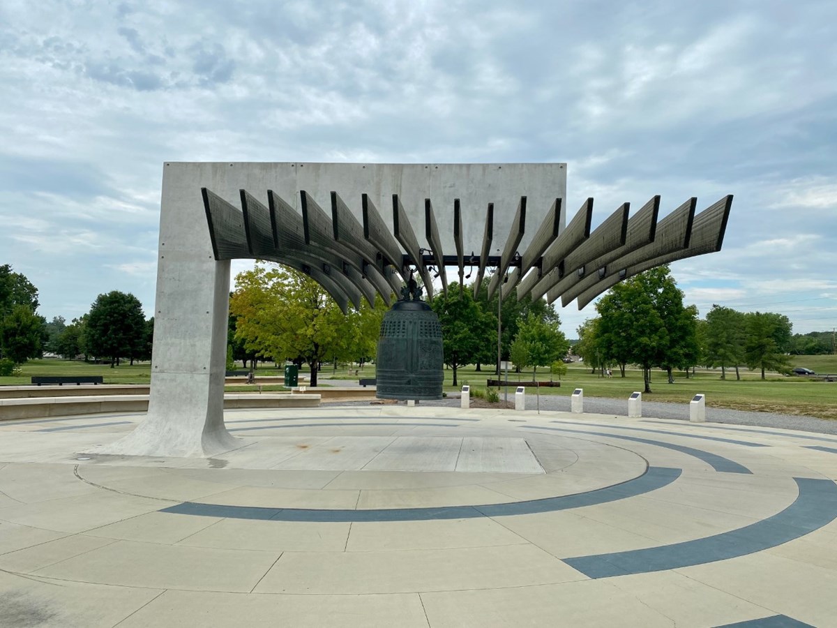 Photo of large bronze bell hanging from concrete structure. The ground is a circular, concrete walkway in an outdoor park.
