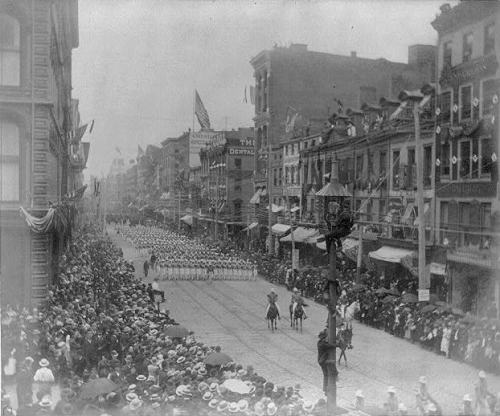 Black and white photo of a funeral procession going down the street with crowds of people on the sidewalk on either end and high buildings surrounding